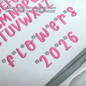 May include: Close-up of embroidered text on white fabric. The word "flowers" and the numbers "2026" are in pink, with small gray flower accents. The top of the image shows the alphabet in pink. The Etsy shop URLs are visible at the top.