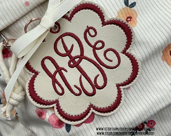 May include: A cream-colored, flower-shaped keychain tag with a burgundy border and embroidered initials. A cream ribbon and a rose gold keyring are attached. The background is a floral fabric with a cream ribbed texture.