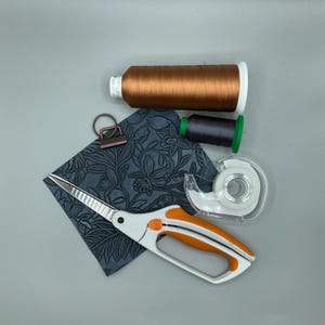 May include: An overhead shot of craft supplies on a gray surface. Includes a spool of brown thread, a spool of dark gray thread, a pair of white and orange scissors, a roll of tape, a metal clip, and a square of embossed blue leather.