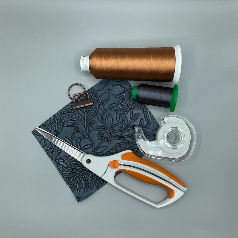 May include: An overhead shot of craft supplies on a gray surface. Includes a spool of brown thread, a spool of dark gray thread, a pair of white and orange scissors, a roll of tape, a metal clip, and a square of embossed blue leather.