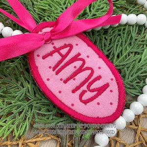 May include: A pink, oval-shaped embroidered tag with the name "Amy" in a darker pink font. The tag has a dotted pattern and a bright pink border. A bright pink ribbon is attached to the tag. The tag is displayed on a bed of greenery and white beads.