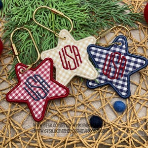 May include: Three star-shaped keychains with gold-tone metal loops. One features a red and white gingham pattern with blue USA embroidery, one is beige gingham with red USA embroidery, and one is navy blue and white gingham with red USA embroidery.