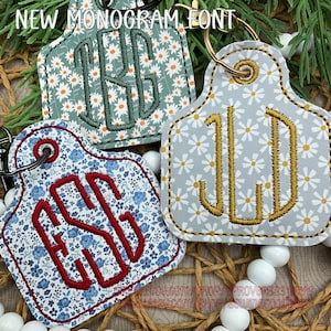 May include: Three keyrings with embroidered monograms. One features a blue floral pattern with red letters, another has a green floral design with dark green letters, and the third is grey with gold letters. Each has a metal ring.