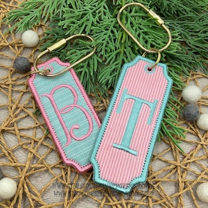 May include: Two rectangular keychains with rounded corners and gold-tone metal rings. One keychain is light blue with white pinstripes and a pink embroidered letter B. The other is pink with white pinstripes and a light blue embroidered letter T.