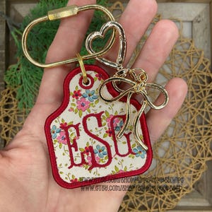 May include: A keychain featuring a cream-colored fabric tag with a floral pattern in pink, blue, and green, embroidered with the red letters ESG. The tag has a red border and is attached to a gold-toned metal ring with a heart-shaped clasp and bow.