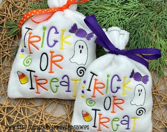 Op de afbeelding: Twee witte Halloween-traktatiezakjes met geborduurde tekst en afbeeldingen. De zakken lezen "Trick or Treat" in kleurrijke letters, met afbeeldingen van een spook, snoep en krullen. Eén zak heeft een oranje stippenlint, de andere een paars lint.
