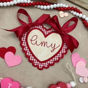 May include: A heart-shaped decoration with a red and white gingham border and a cream-colored center. The word "Amy" is embroidered in red script. It has a red satin ribbon and is surrounded by felt hearts and beads.