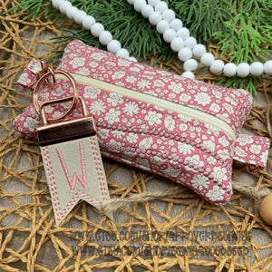 May include: A pink floral print zippered pouch with a matching keychain. The keychain has a light-colored leather tag with the letter "W" stitched in pink. The pouch and keychain are on a woven surface.