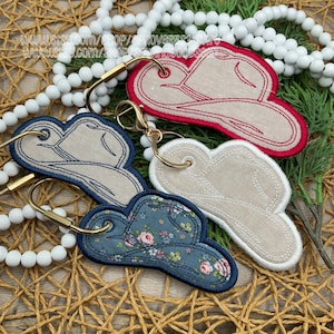 May include: Embroidered cowboy hat keyrings in various colours. One is red with a beige centre, another is blue with a floral pattern, and two are beige. Each keyring has a gold-coloured clasp and ring.