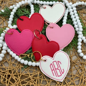 May include: A collection of heart-shaped keychains in various colors, including red, pink, and white. Some keychains have embroidered initials. Each keychain has a metal ring and clasp for attachment.