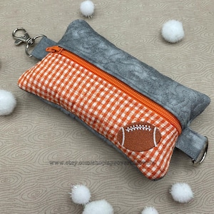 May include: A small orange and gray zippered pouch with a football design. The pouch has a checkered pattern and a zipper pull with a key ring.