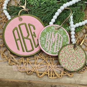 May include: Three round keychains with gold-colored hardware. The largest keychain is pink with green embroidered initials. The middle keychain is green with white floral embroidery and initials. The smallest keychain has a floral pattern.