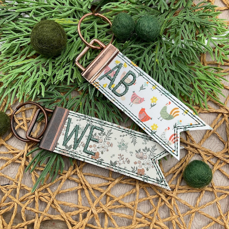 May include: Two keychains with floral and chicken patterns. One keychain has the letters "AB" embroidered in green, the other has "WE". Both have metal hardware and a decorative stitched edge. Green felt balls and greenery are in the background.