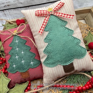 Op de afbeelding: Twee rechthoekige kussens met kerstthema. Eén is rood met witte strepen en een groene vilten kerstboom, de andere is crèmekleurig met rode strepen en een groene vilten boom. Beide hebben decoratieve strikken.