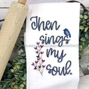 Könnte beinhalten: Weißes Küchentuch mit der Stickerei "Then sings my soul". Ein blauer Vogel und florale Akzente sind ebenfalls auf dem Tuch gestickt. Auf der linken Seite des Tuchs liegt ein Nudelholz aus Holz.