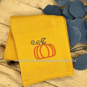 May include: Folded yellow waffle-weave towel with an embroidered orange pumpkin design. The pumpkin has a dark gray stem and vines. The towel is folded and resting on a wooden surface. The Etsy shop URL is visible.