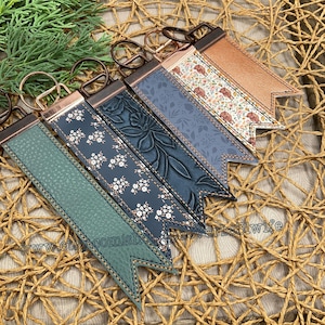 May include: A collection of keychains with various designs. The keychains feature leather straps in shades of green, blue, and brown, adorned with floral patterns and embossed textures. Each keychain has a metal ring and a decorative end.