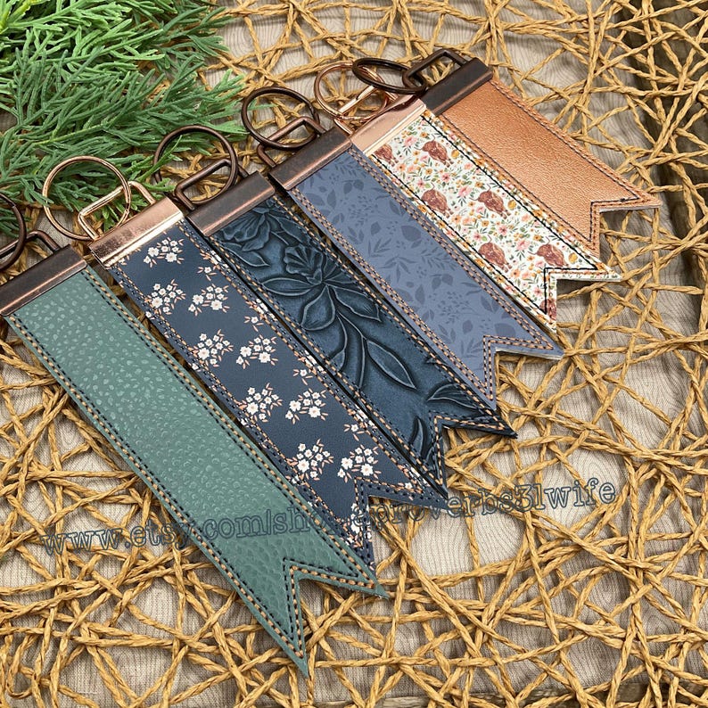 May include: A collection of keychains with various designs. The keychains feature leather straps in shades of green, blue, and brown, adorned with floral patterns and embossed textures. Each keychain has a metal ring and a decorative end.