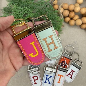 May include: A collection of personalized keychains with gold-tone hardware. The keychains feature various colors, including pink, mint green, orange, and white, each embroidered with a different letter in a contrasting color. The letters include J, H, P, K, T, and A.