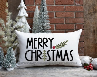 Puede incluir: Almohada blanca con texto negro que dice "Feliz Navidad" con una rama de acebo verde y roja. La almohada está sobre una superficie de madera con pequeños árboles de Navidad artificiales blancos y verdes.