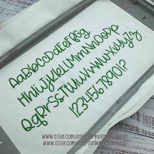 Op de afbeelding: Witte stof met groene geborduurde cursieve letters, cijfers en symbolen. De stof wordt in een metalen borduurring gehouden. Etsy shop URL's zijn onderaan zichtbaar.