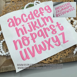 Op de afbeelding: Een witte stof met het kleine alfabet geborduurd in een helder roze, cursieve lettertype. De letters zijn in rijen gerangschikt. Een kleinere afbeelding in de rechterbovenhoek toont het hoofdletteralfabet in hetzelfde lettertype. Etsy shop-URL's staan onderaan afgedrukt.
