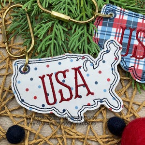 May include: A white keychain shaped like the map of the USA with red and blue polka dots and the text USA embroidered in red thread. A second keychain with a red, white, and blue plaid pattern is partially visible, both attached to gold rings.