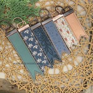 May include: A collection of six keychains with decorative leather straps in various colors and patterns. The straps include floral, embossed, and solid designs, each with a metal clasp and ring. The keychains are arranged on a woven surface.