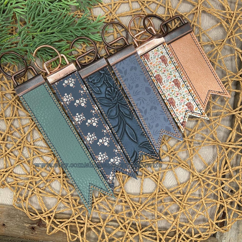 May include: A collection of six keychains with decorative leather straps in various colors and patterns. The straps include floral, embossed, and solid designs, each with a metal clasp and ring. The keychains are arranged on a woven surface.
