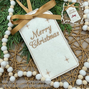 May include: A white felt Christmas gift tag with gold embroidered text reading "Merry Christmas." The tag has a gold ribbon and star accents. The tag is surrounded by decorative beads and greenery.