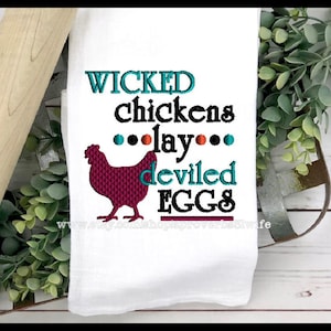 May include: White kitchen towel with the embroidered text "WICKED chickens lay deviled EGGS" in teal, black, and burgundy. A burgundy chicken silhouette is also embroidered on the towel. The towel is displayed with a rolling pin and greenery.
