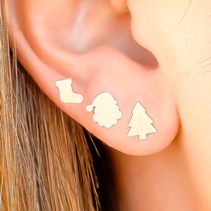May include: A close-up of an ear with three small, gold-colored Christmas-themed stud earrings. The earrings are shaped like a stocking, Santa hat, and Christmas tree. The ear is set against a blurred background.