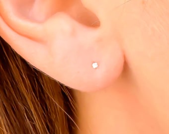 May include: A tiny, round, silver-coloured stud earring is centred on the earlobe. The earring features a small, reflective metallic surface that catches the light, creating a subtle point of brightness against the skin of the earlobe.