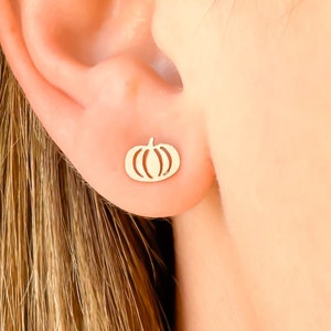 May include: Gold-colored pumpkin stud earrings. The earrings are small, with a simple outline of a pumpkin shape. The earrings are worn on a person's ear.