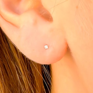 May include: A close-up of a person's ear with a small, round, clear stud earring. The earring has a single, sparkling, clear stone. The person's hair is a light brown color.