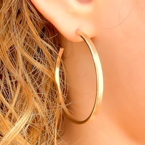 May include: A close-up of a gold hoop earring. The earring is a simple, circular design with a smooth, polished surface. The earring is worn on the ear, with the hair in the background.