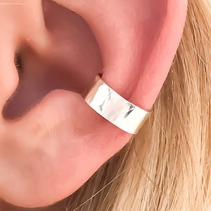 May include: A polished silver-tone ear cuff with a smooth, wide band design. The metal accessory is positioned securely on the outer cartilage of the ear, featuring a reflective, high-shine finish that catches the light against the skin.