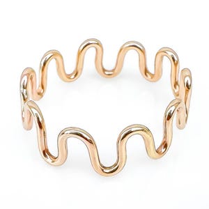 May include: A gold-colored ring with a wavy, continuous design. The ring's shape is a series of connected curves, creating a unique and modern aesthetic. The ring is set against a white background, highlighting its metallic sheen.