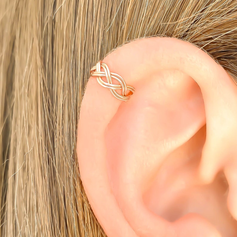 May include: A rose gold-colored ear cuff featuring a delicate, interwoven braided wire design. The metal band wraps around the outer edge of the ear cartilage, showcasing a continuous, symmetrical pattern of overlapping metallic loops.
