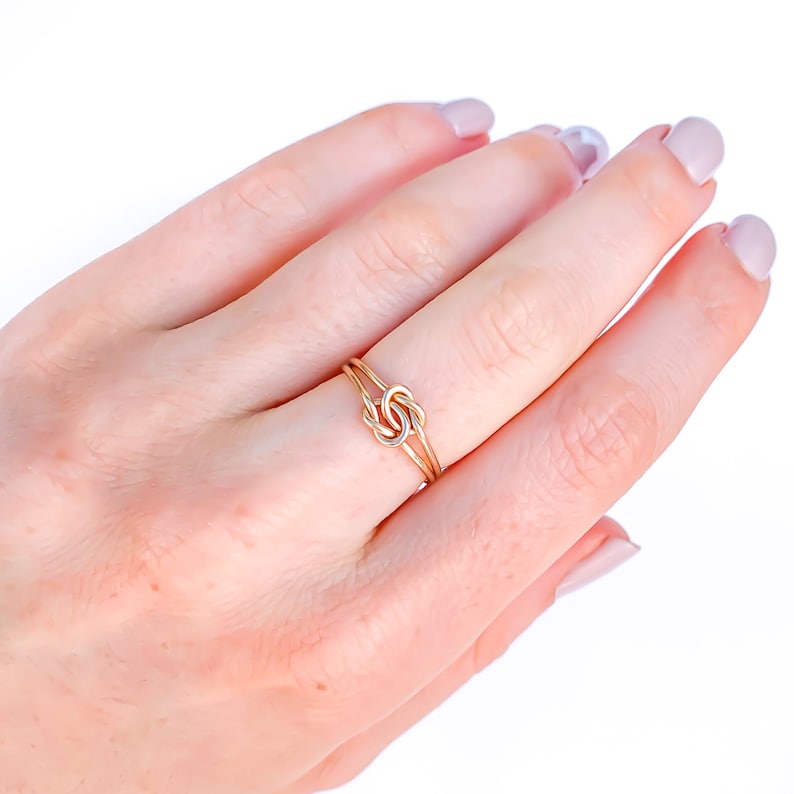May include: A delicate ring featuring a central knot design crafted from thin, polished gold-toned wire. The band consists of two slender, parallel wires that converge into a small, intricate knot at the centre, creating a refined and minimalist appearance.