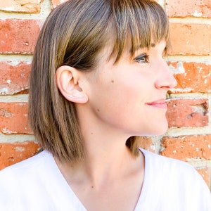 May include: A woman with brown hair wearing a white shirt and a small silver ear cuff.