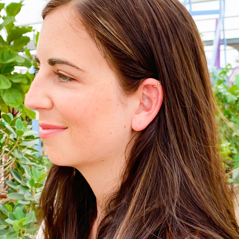 May include: A gold-colored ear cuff is worn on the upper ear of a person. The ear cuff has a delicate, openwork design. The person has long, brown hair and is smiling. The background is blurred with green foliage.