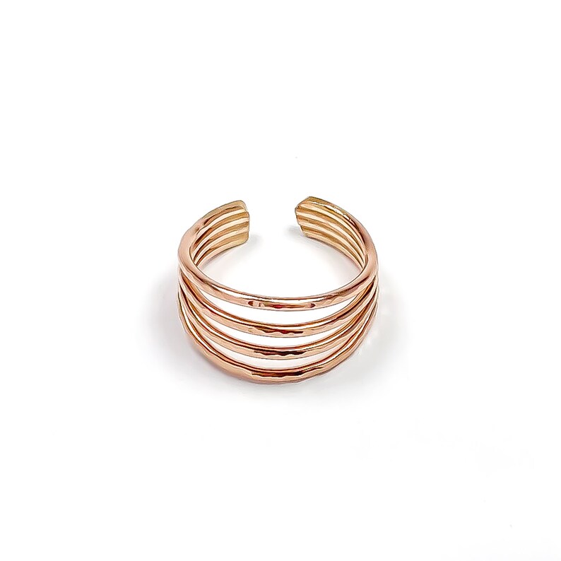 May include: A rose gold-toned open ring featuring a multi-band design composed of five thin, parallel wires. The metal surface displays a subtle hammered texture, creating a reflective finish across the stacked, circular structure.