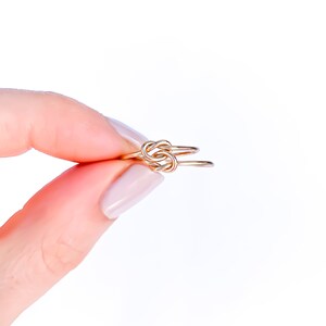 May include: A thin gold ring featuring a central knot design. The band is constructed from a smooth, polished gold-toned metal wire that loops over itself to form a symmetrical knot at the front, creating a delicate and continuous circular shape.