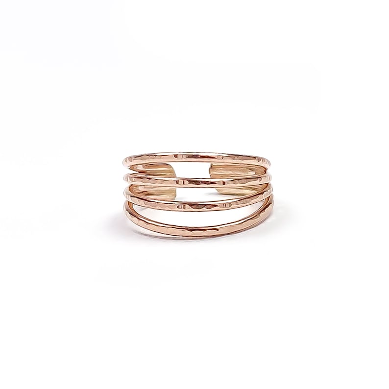 May include: A rose gold ring with a multi-band design. The ring features four hammered bands, each with a slightly different curve, creating a layered effect. The open design allows for comfortable wear. The ring is set against a white background.