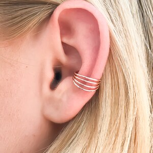 May include: A gold-colored ear cuff with a layered design is shown on an ear. The cuff has three parallel bands, each with a textured surface. The ear and surrounding hair are in soft focus, providing a natural backdrop.