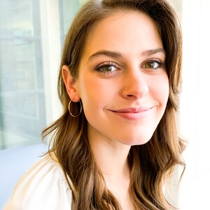 May include: A woman with long brown hair wearing a white shirt and gold hoop earrings. She is smiling and looking at the camera.