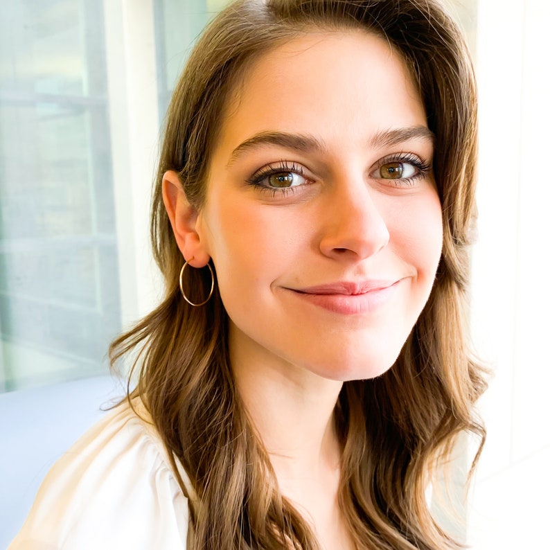 May include: A woman with long brown hair wearing a white shirt and gold hoop earrings. She is smiling and looking at the camera.