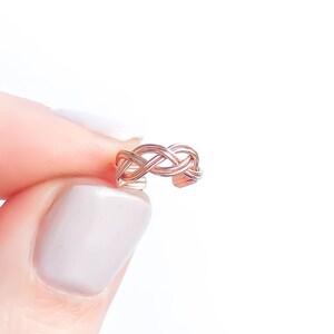 May include: A rose gold ear cuff with a woven design. The ear cuff is held between fingers against a white background. The cuff's intricate pattern is visible, showcasing its delicate craftsmanship. This piece of jewelry is designed to be worn on the ear.