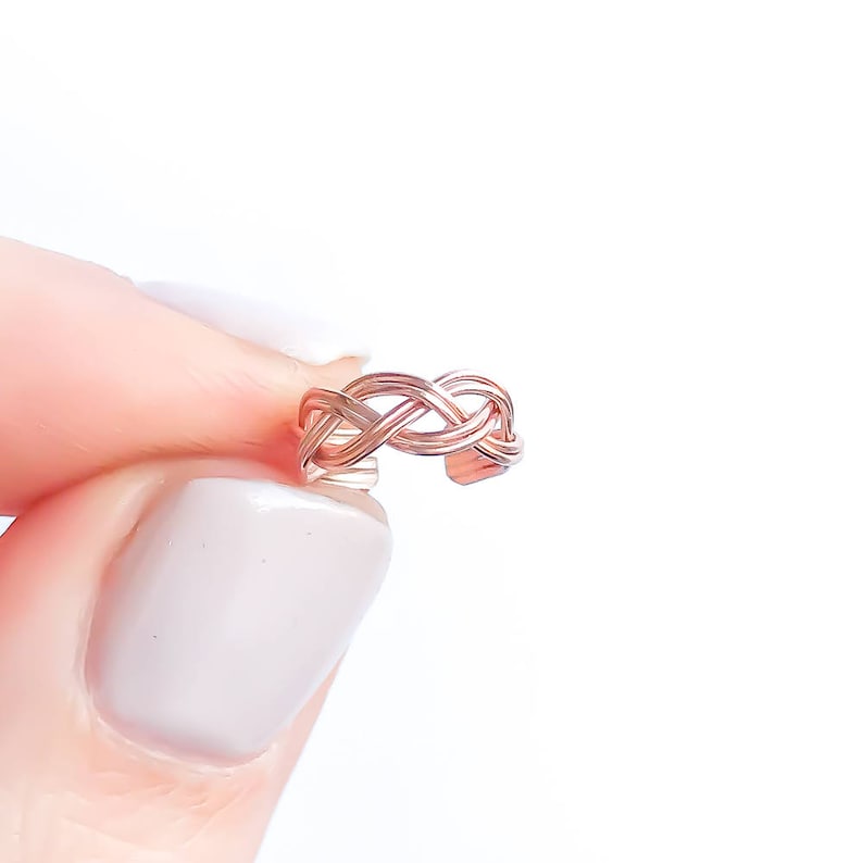 May include: A rose gold ear cuff with a woven design. The ear cuff is held between fingers against a white background. The cuff's intricate pattern is visible, showcasing its delicate craftsmanship. This piece of jewelry is designed to be worn on the ear.